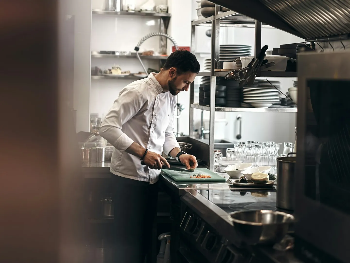 Cuisinier en veste blanche et tablier gris découpant des légumes sur une planche verte, dans une cuisine professionnelle en inox
