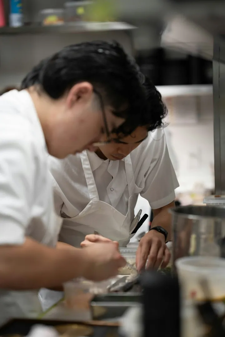 Deux jeunes cuisiniers en tenue blanche, dont l'un porte un tablier blanc, penchés sur le dressage d'une préparation en cuisine